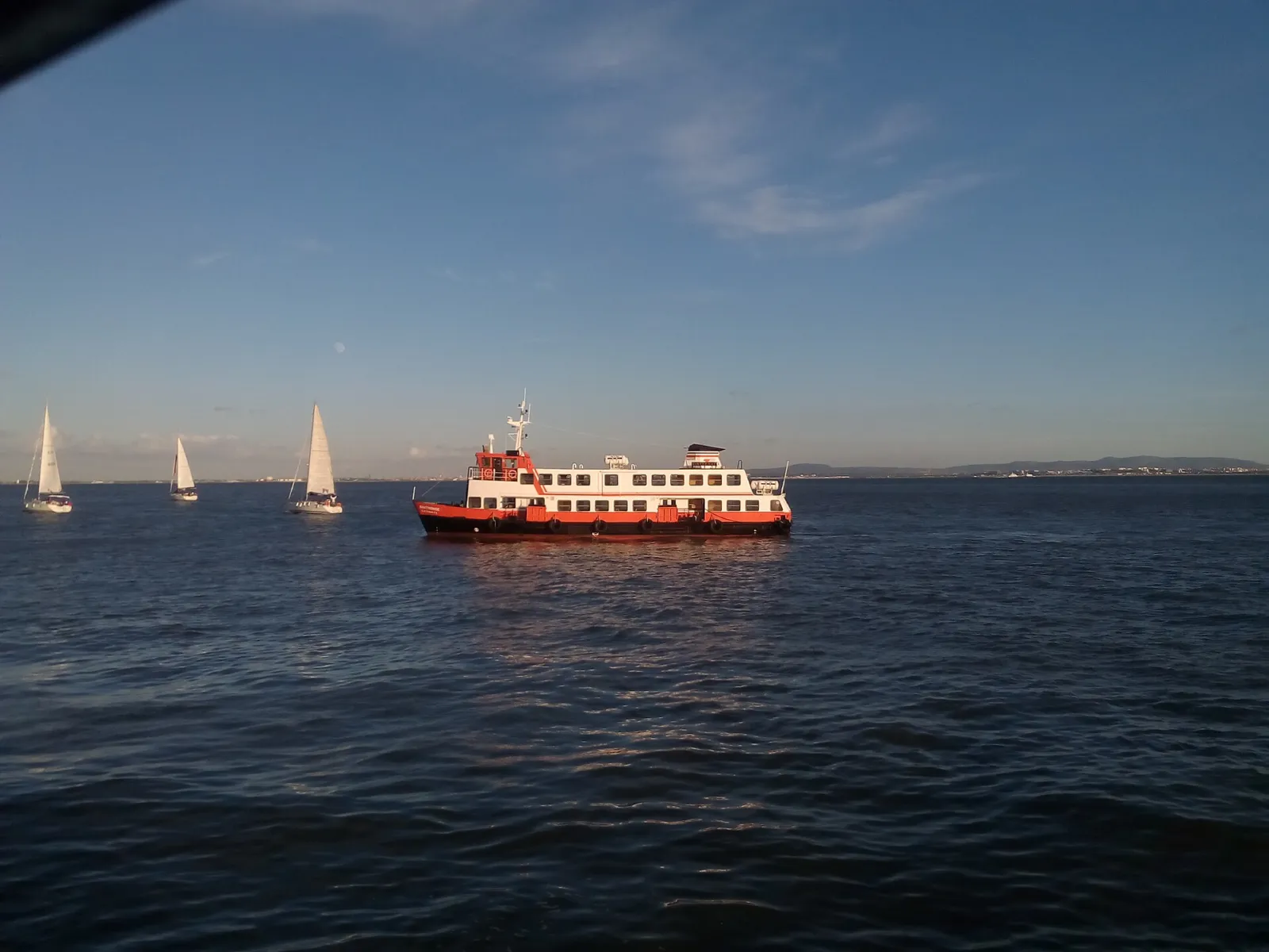 Ferry de pasajeros rojo y blanco navegando por el río Tajo con veleros al fondo